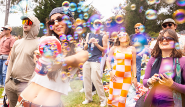 A group of people outdoors enjoy bubbles, with a woman in front playfully spraying bubbles from a can, while others smile and watch.