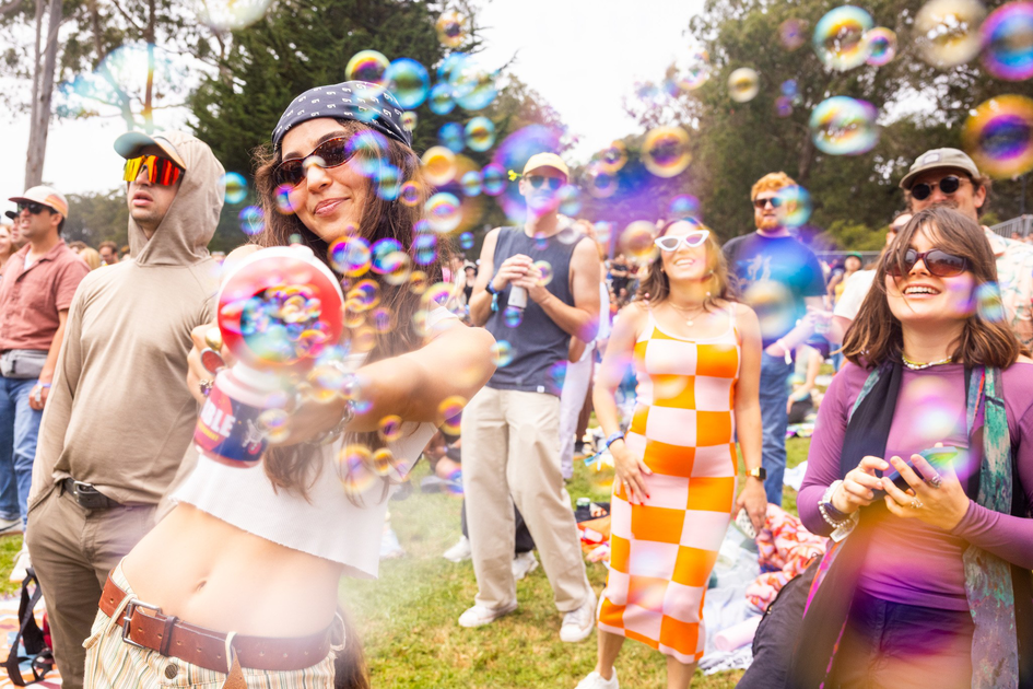 A group of people outdoors enjoy bubbles, with a woman in front playfully spraying bubbles from a can, while others smile and watch.