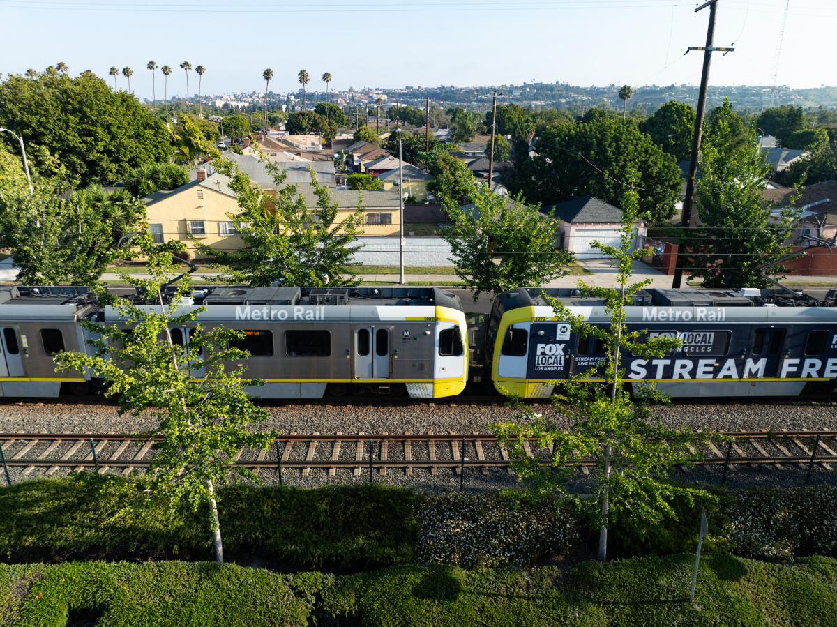 A light-rail metro train in Los Angeles passes through a neighborhood with houses and trees.