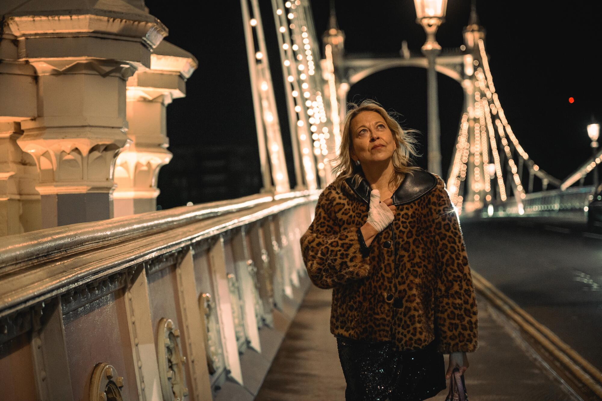 A woman in a leopard-print coat walking down a pathway on a bridge in the night time.