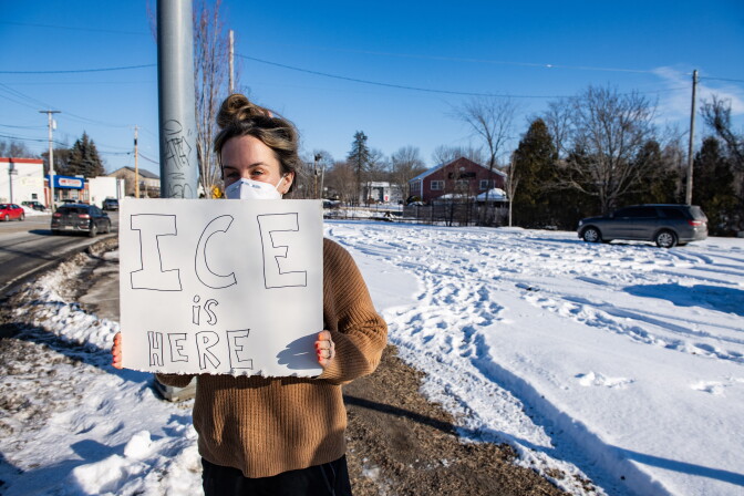 A person holds an anti-ICE sign. Footprints are visible in the snow near her.