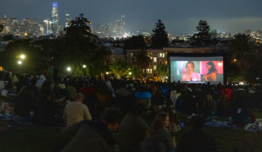 A large crowd sits on a grassy hill watching a movie on an outdoor screen at night, with a city skyline lit up in the background.