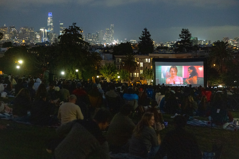 A large crowd sits on a grassy hill watching a movie on an outdoor screen at night, with a city skyline lit up in the background.