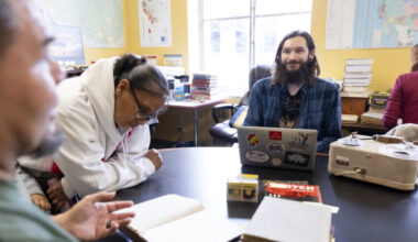 Berkeley graduate student Tyler Lee-Wynant sits smiling at a table inside the California Language Archive, surrounded by archival materials, a laptop, an audio recording player while speaking with tribal visitors.