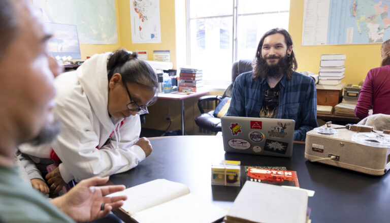 Berkeley graduate student Tyler Lee-Wynant sits smiling at a table inside the California Language Archive, surrounded by archival materials, a laptop, an audio recording player while speaking with tribal visitors.