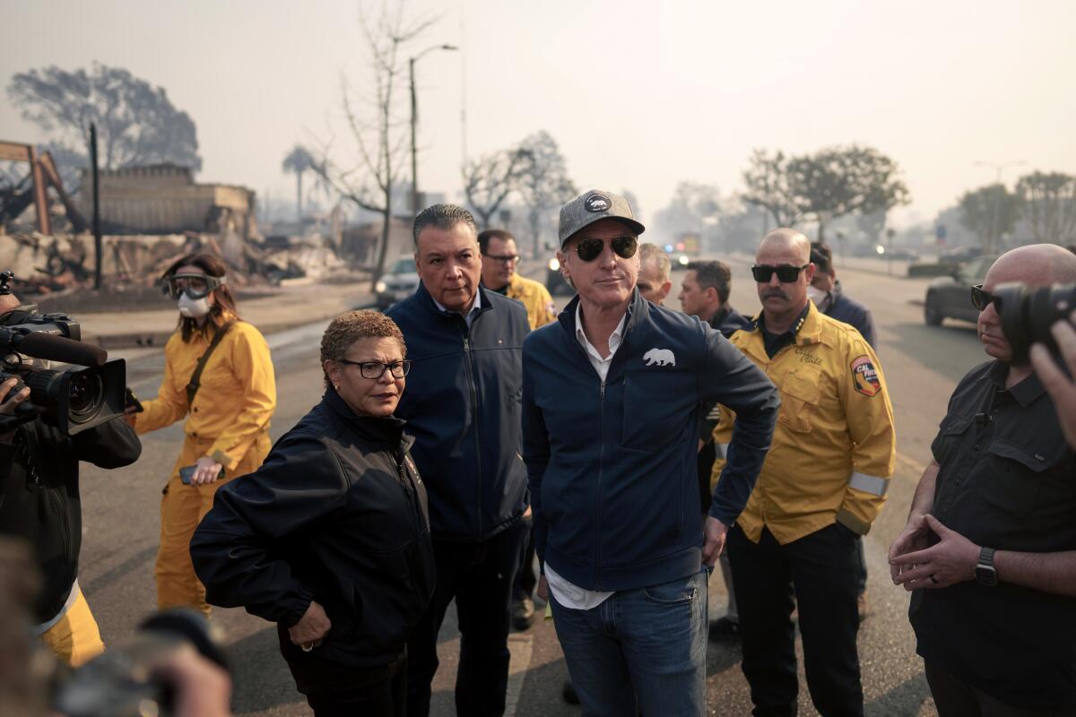 Karen Bass stands alongside Gavin Newsom on a city street. In the background are burned buildings.