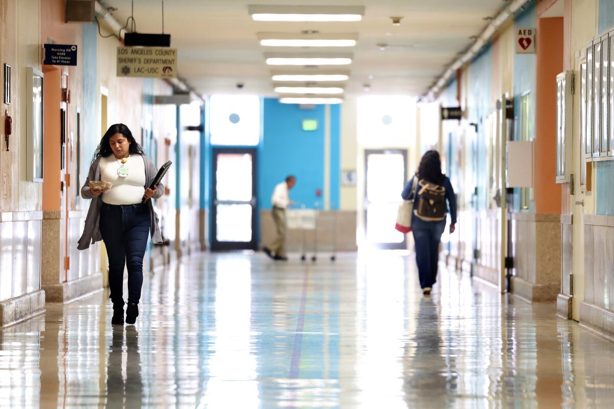people walk down a hallway in a hospital, one woman looks at her phone