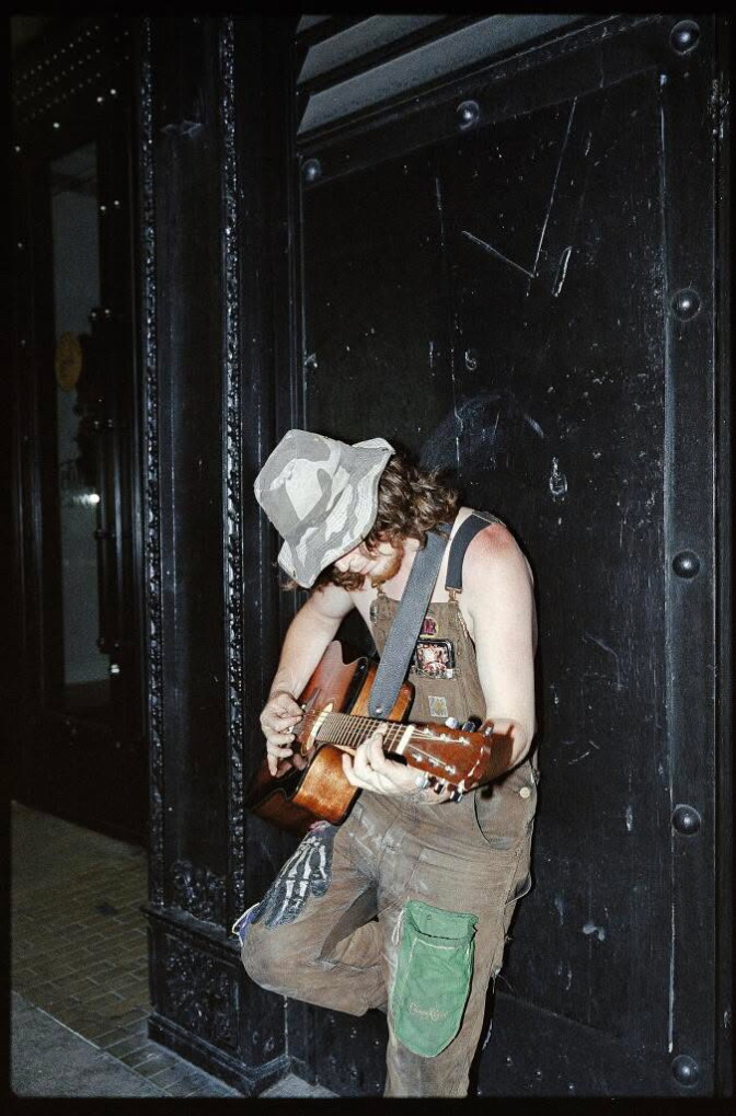 A man in overalls and a camouflage hat plays guitar against a black wall.