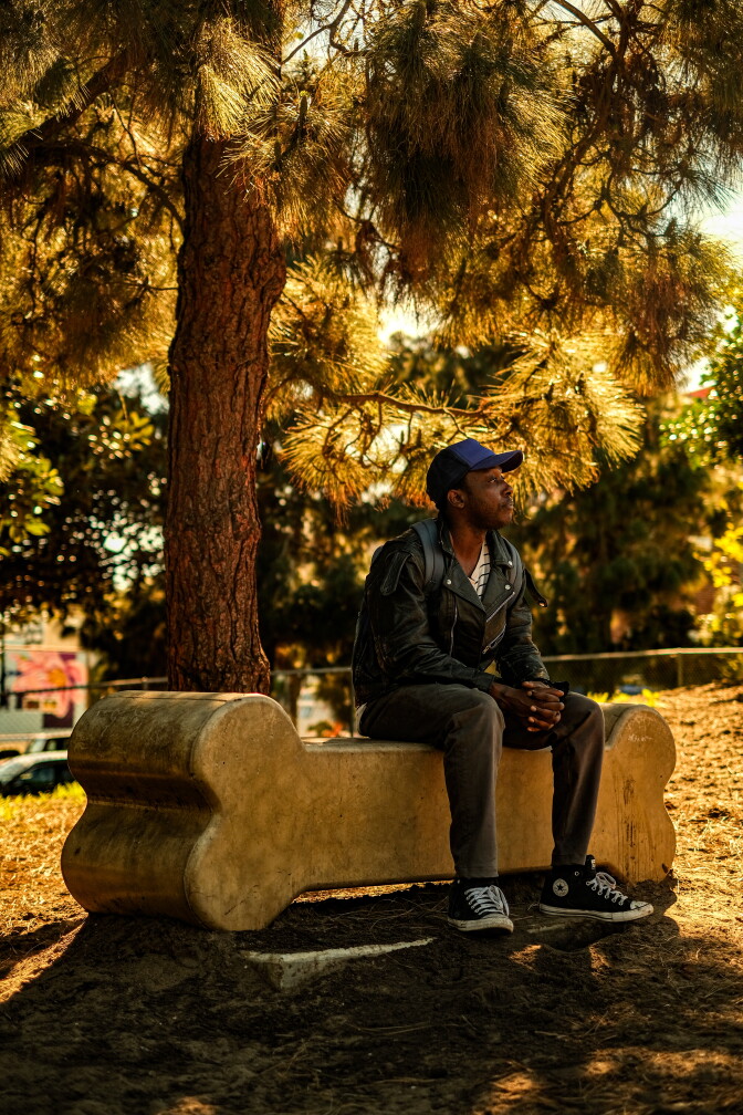 A man sits on a park bench shaped like a dog bone. He is sitting under a tree and looking off into the distance.