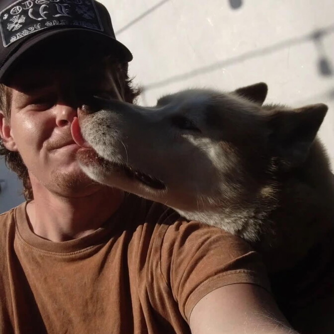 A dog licks a mans face. The face is partially shaded by a black hat.