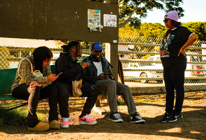 Three people sit on a green bench smiling and talking. A fourth person stands looking at the bench.