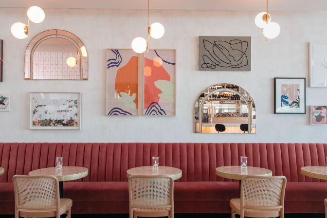 The interior of a restaurant, which has red banquet seats with four marble topped tables in front of them. On the wall is an assortment of abstract paintings and mirrors that have a rainbow shaped top.