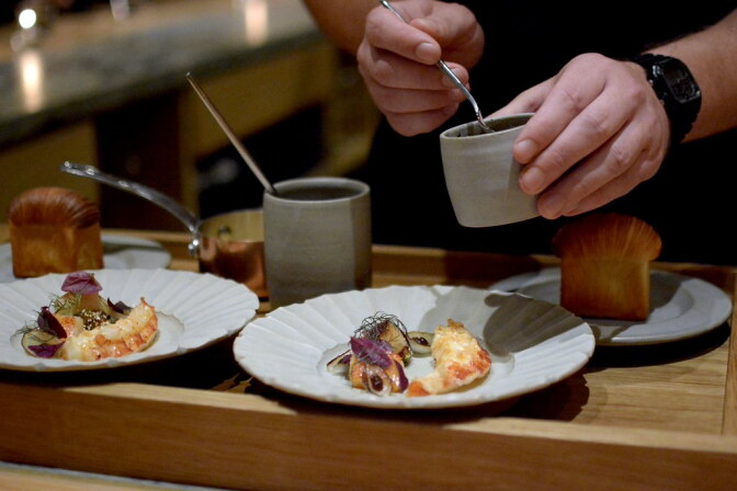 Two light skinned hands are holding a small grey container with a teaspoon inside. They're about to pour something over the waiting white dishes below which hold two lobster pieces covered with a garnish.