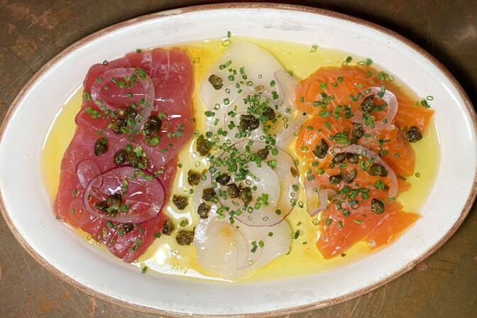 A white oval plate contains three different piles of raw fish; one is dark red, one is orange and one is white; they're sitting in a bed of olive oil, onions and capers.