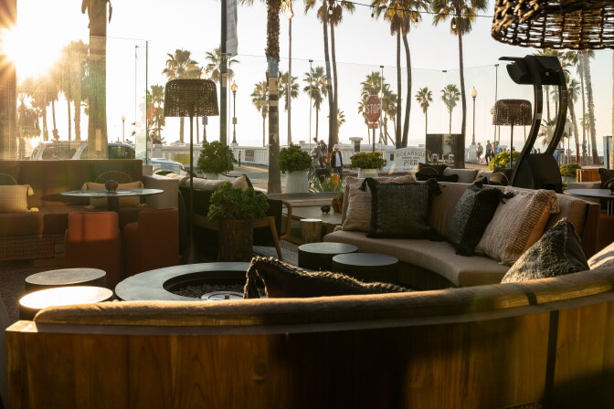 The interior of a restaurant lounge has a round brown sofa with stools and a firepit in front of it; it sits next to a glass barrier, through which you can see the beach with large palm trees outlined in the sun.