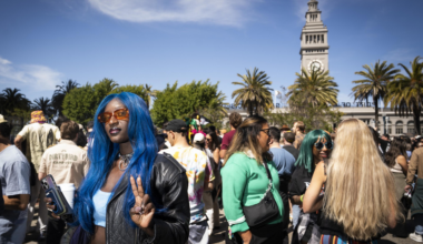 A person with long blue hair and orange sunglasses flashes a peace sign at a crowded outdoor event. A clock tower and palm trees are visible in the background.