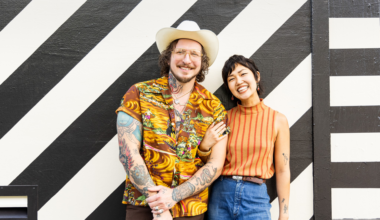 A smiling man in a white hat and colorful shirt stands next to a laughing woman in an orange striped top and blue jeans, both with tattoos, in front of a black-and-white striped wall.