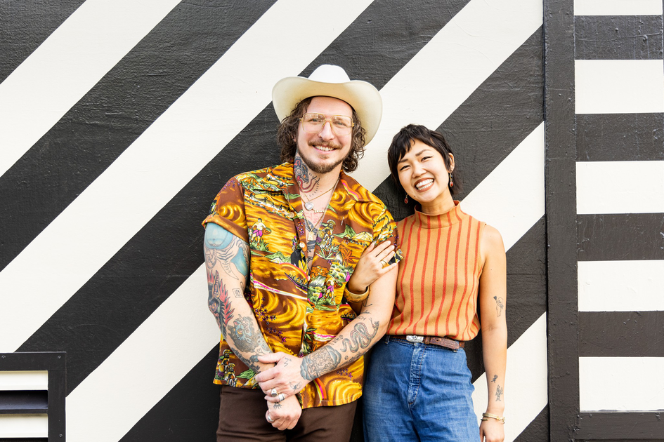 A smiling man in a white hat and colorful shirt stands next to a laughing woman in an orange striped top and blue jeans, both with tattoos, in front of a black-and-white striped wall.