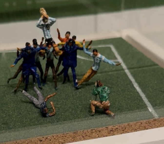 A tiny scene of miniatures on a soccer field. A soccer player is hoisted above the heads of other players.