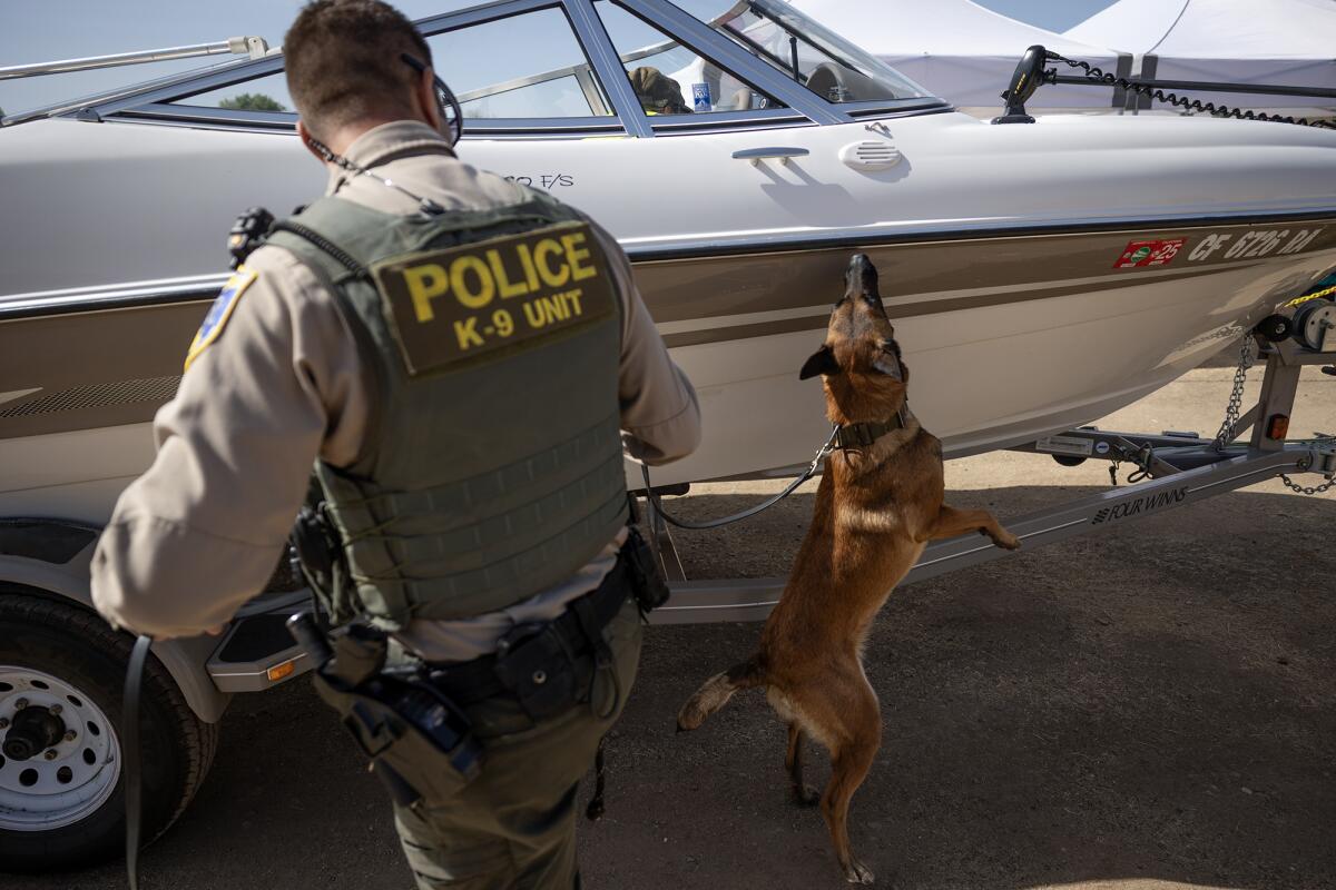 A law enforcement officer wearing a vest labeled "POLICE K-9 UNIT" stands beside a trailer-mounted speedboat 