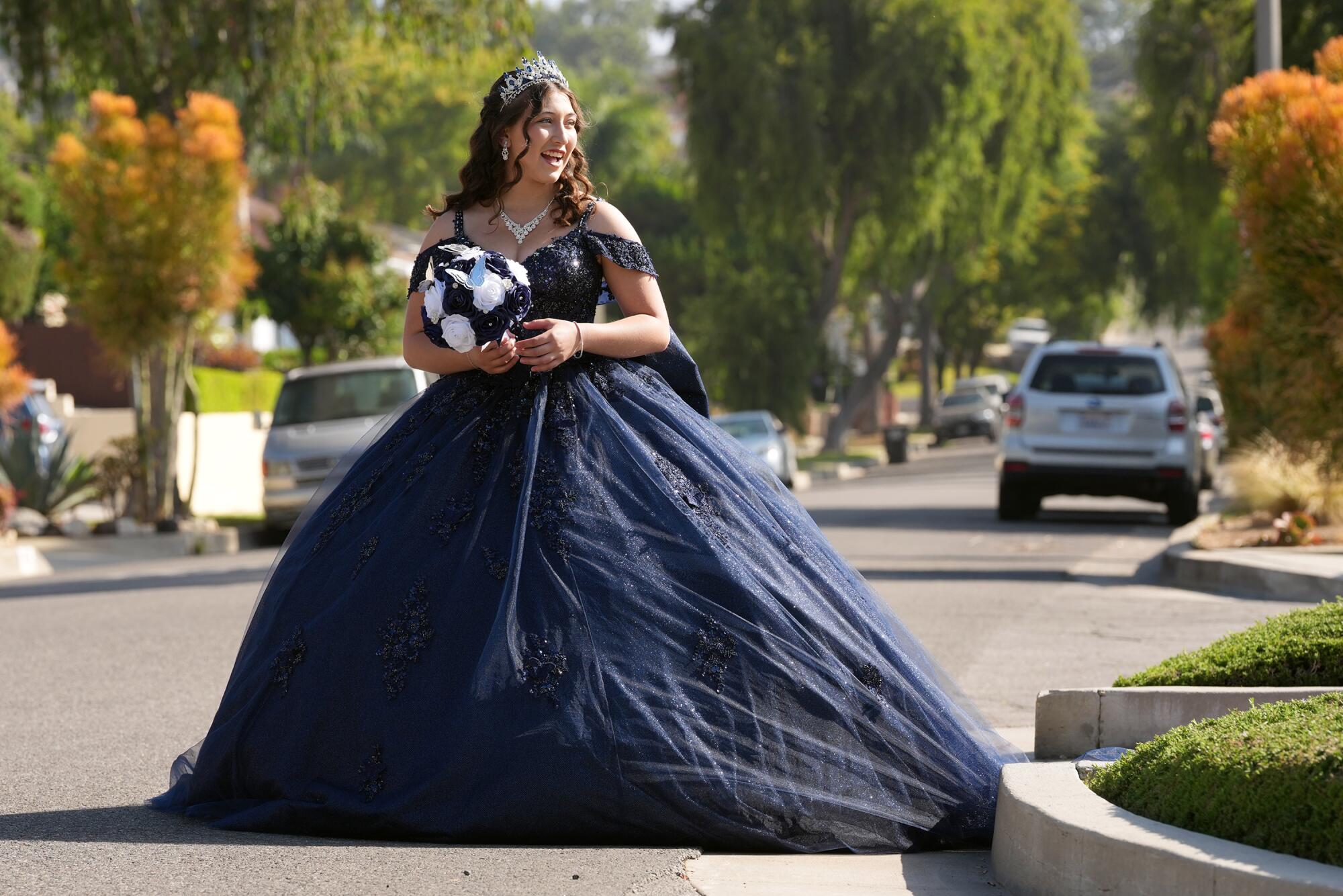 A teen in a navy blue dress, sparkly necklace and tiara holds a white bouquet where a street meets a park.