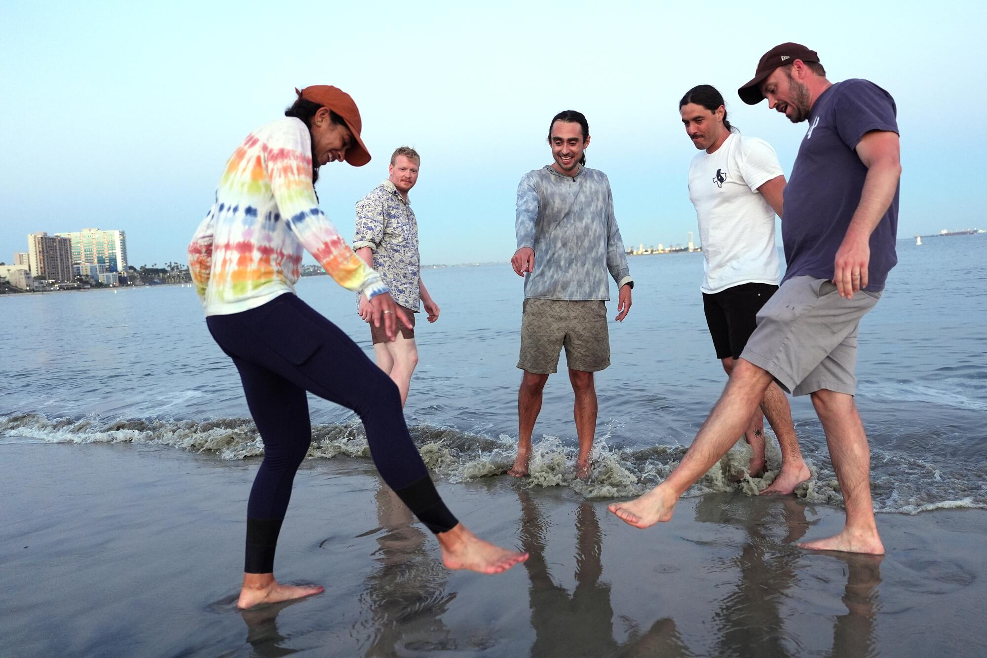 Five people dip their toes in the water, pointing out one of their sock tans. 