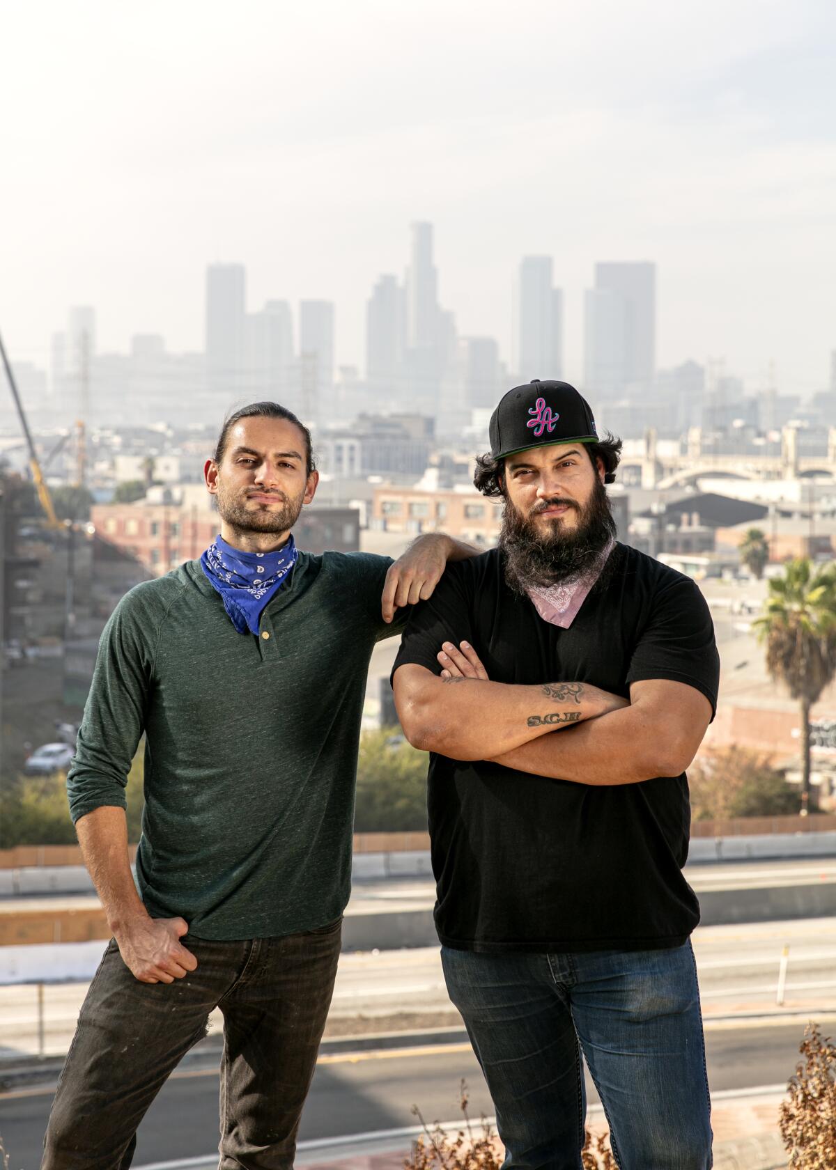 A man in a dark blue shirt standing next to another man, in a dark cap and short-sleeved shirt, arms crossed