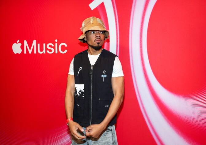 A young Black man with a mustache and goatee wearing jeans, a white t-shirt, black vest and patchwork khaki bucket hat. He's looking off to his left with a red and white background behind him with the Apple Music logo on it.