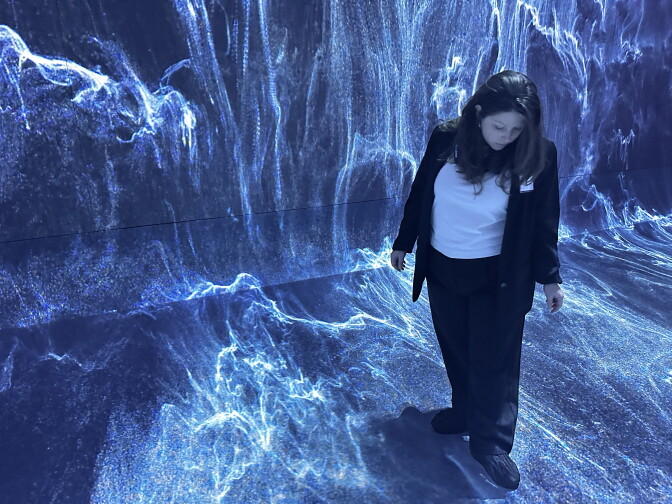 A woman stands in a room with an abstract light blue, dark blue and white image projected across the floors and wall.