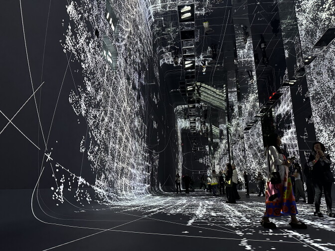 A room with curved walls on its left side and mirrored pillars on the right, projecting an image of thousands of white dots that form abstract shapes against a black background.
