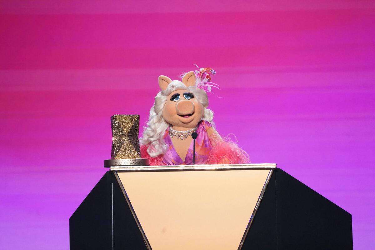 A glammed up Miss Piggy stands at a podium in front of a hot pink backrop