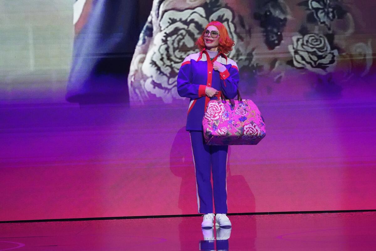 A woman in blue track suit, a red wig and exaggerated makeup stands on a stage.