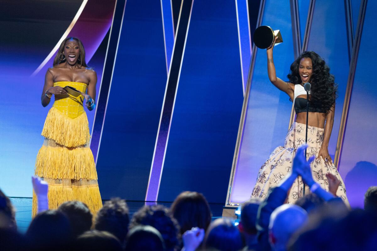 Two women in glam attire -- one holding a trophy up in the air -- face an audience on stage