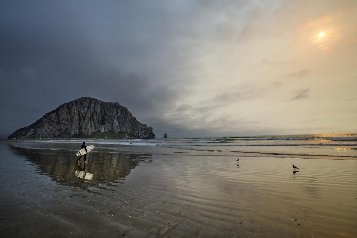 A surfer and Morro Rock are reflected in the water at low tide as the sun goes down with ash visible from a forest fire. 