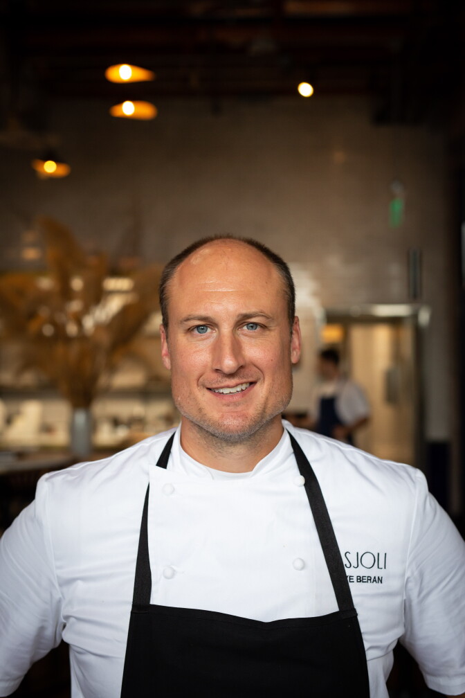A man with a light skin tone and bald head in white chef's coat and black apron standing in restaurant kitchen, smiling at camera.