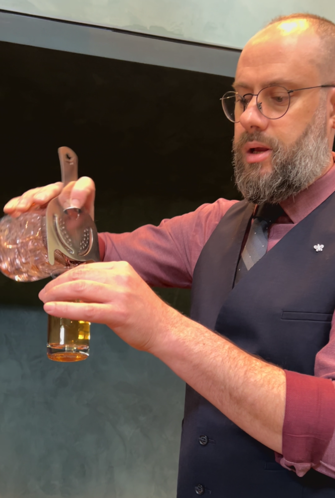 A man with a light skin tone, a shaved head, and a salt-and-pepper beard is wearing thin black wire-framed glasses, a cranberry button-up shirt with sleeves rolled up to his forearms, a navy blue vest, and a grey and blue striped tie. He is holding a metal cocktail shaker, which he is pouring into a clear glass container.