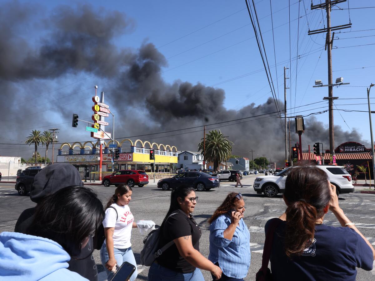 People cross the street as black smoke rises from a blaze inside a cold storage facility in Boyle Heights