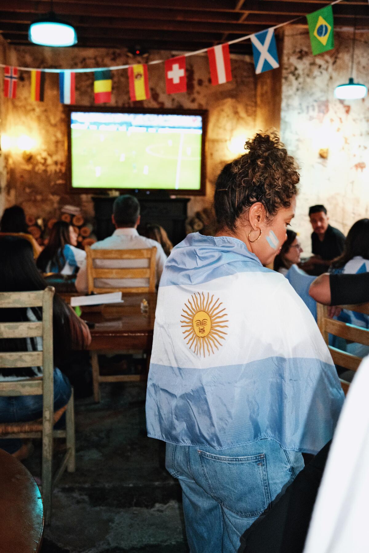A fan wrapped in an Argentina flag watches a futbol game on a screen inside Fuegos LA
