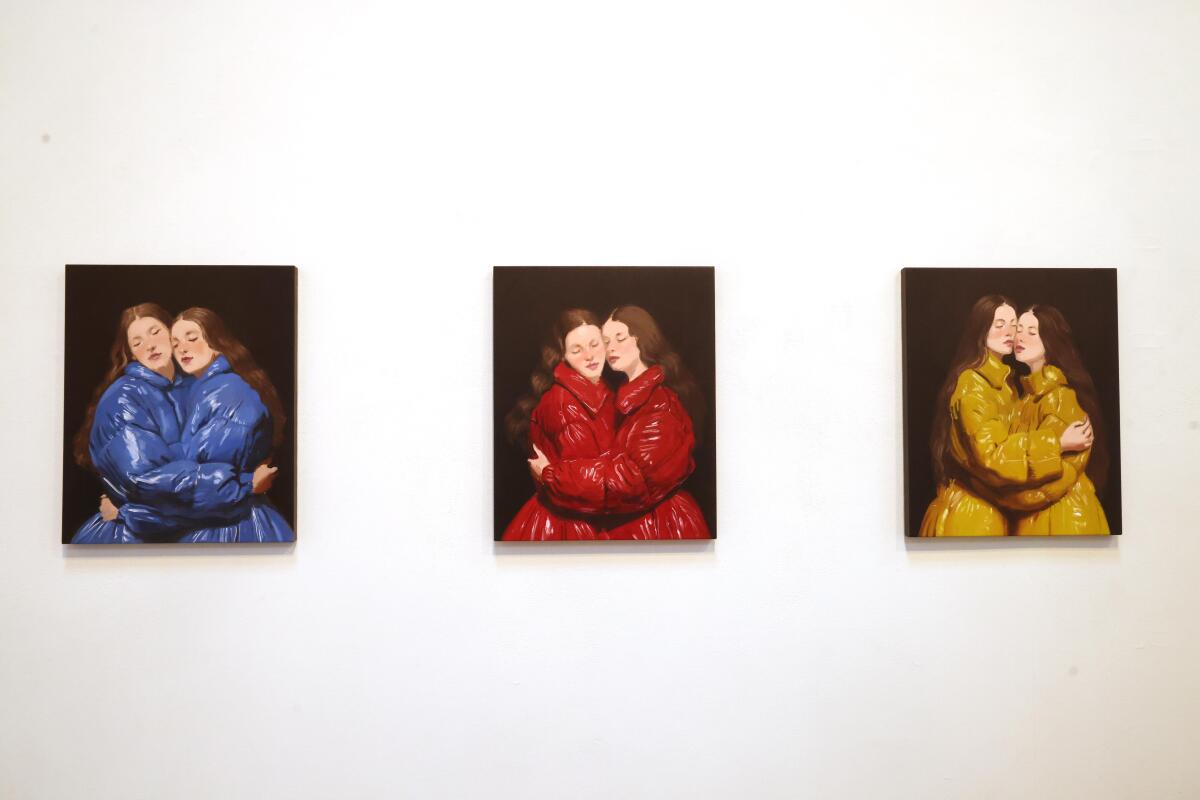 Three paintings of three pairs of women wearing blue, red and yellow puffer jackets hang on a gallery wall. 