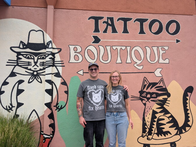 A man and a woman wearing matching cat t-shirts standing in front of a mural of cats.