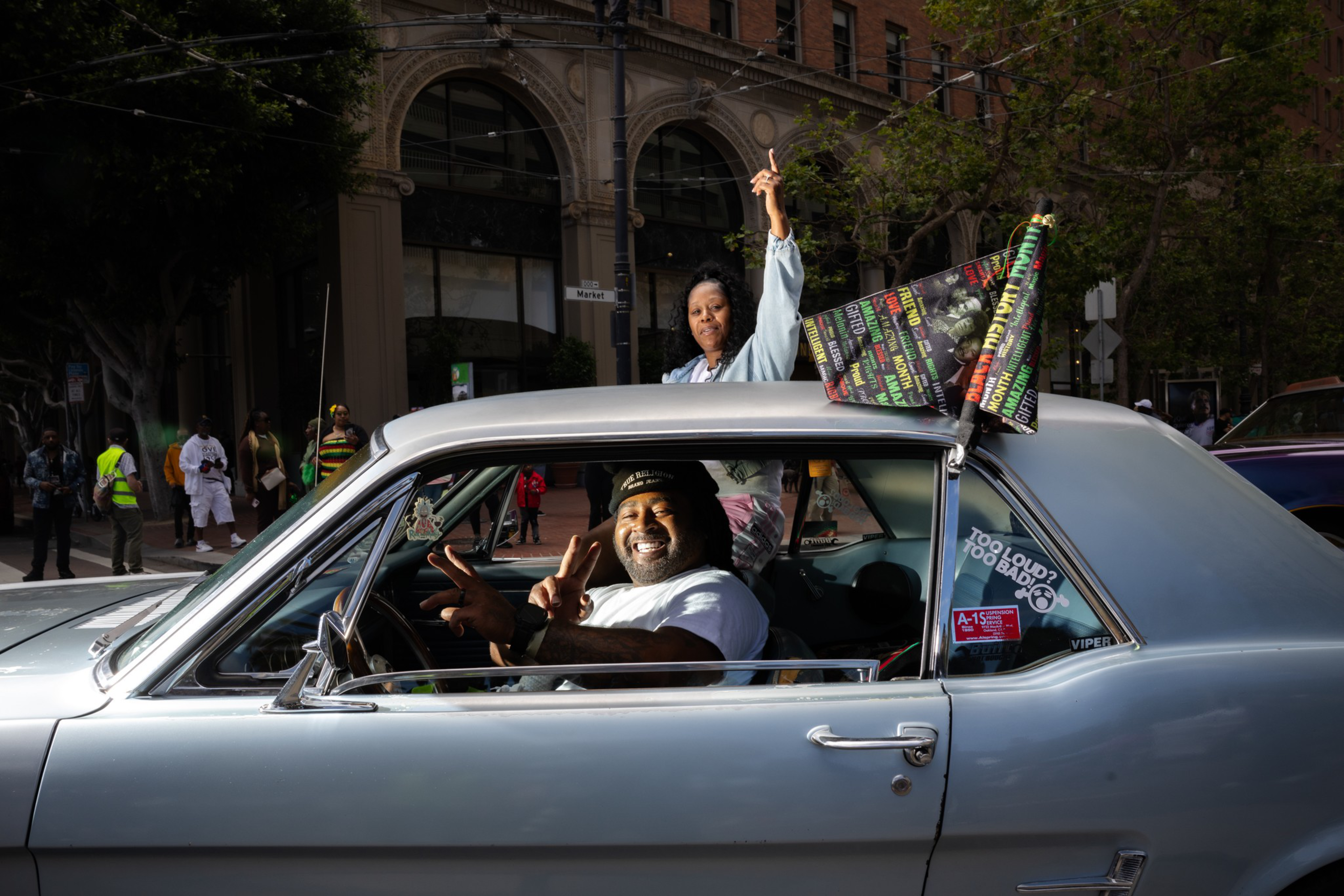A smiling man makes a peace sign while seated in a gray classic car, and a woman stands through the sunroof waving a colorful flag.