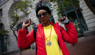 A smiling person wearing sunglasses, a black head wrap with “Juneteenth” decorations, a red jacket, and a yellow shirt with a Juneteenth pendant.