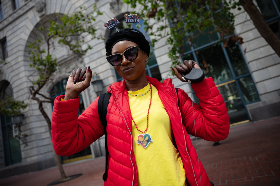 A smiling person wearing sunglasses, a black head wrap with “Juneteenth” decorations, a red jacket, and a yellow shirt with a Juneteenth pendant.