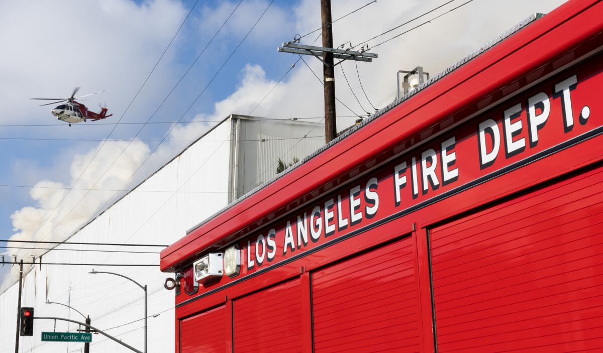 a helicopter flies over a smoking building and an LAFD tanker