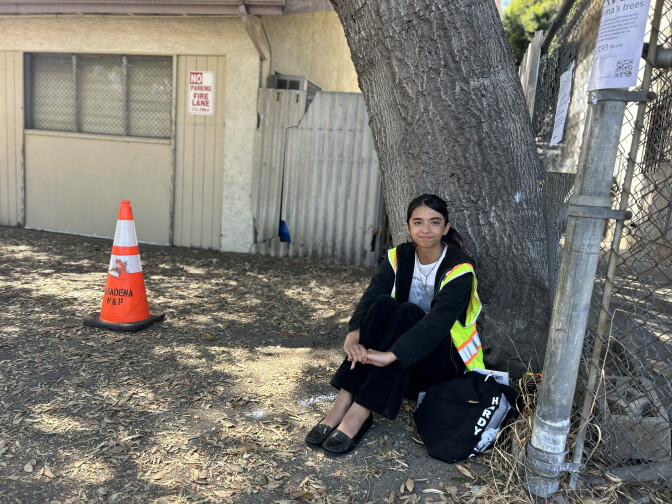 A young woman with light brown skin and dark straight hair wears a safety vest and sits at the base of a tree in a parking lot. 