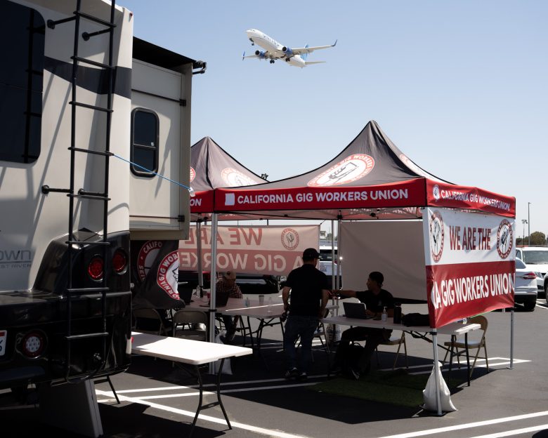 Outdoor scene with a "California Gig Workers Union" tent in a parking lot. Three people sit at tables inside. A plane flies overhead in the clear sky.