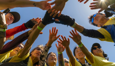 A group of people wearing yellow shirts form a circle, reaching their hands together in the center under a clear blue sky.