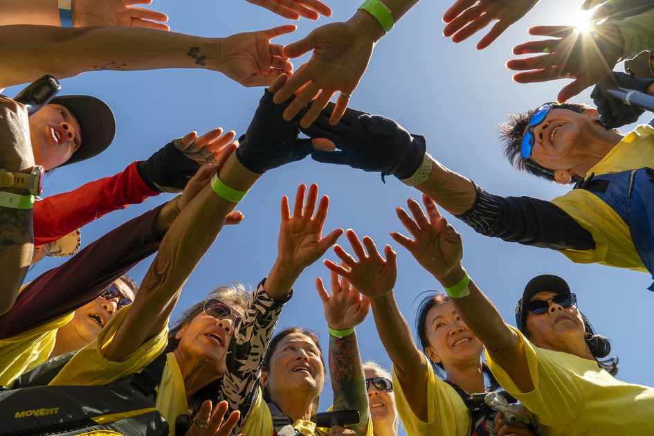 A group of people wearing yellow shirts form a circle, reaching their hands together in the center under a clear blue sky.