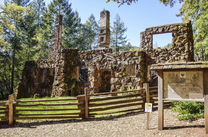 The remnants of an old brick building behind a wooden fence next to tall trees outside. An illustration and description are posted on signage in front of the building.