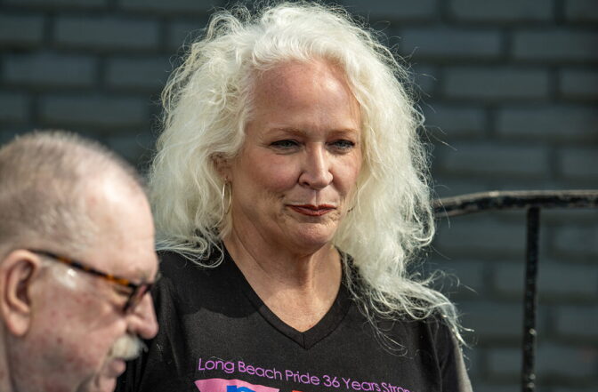 A woman with light skin tone, light blonde hair, wearing a graphic t-shirt, looks out of frame as she stands next to a man with light skin tone, glasses, short hair and a mustache, who is slightly out of focus in the foreground.